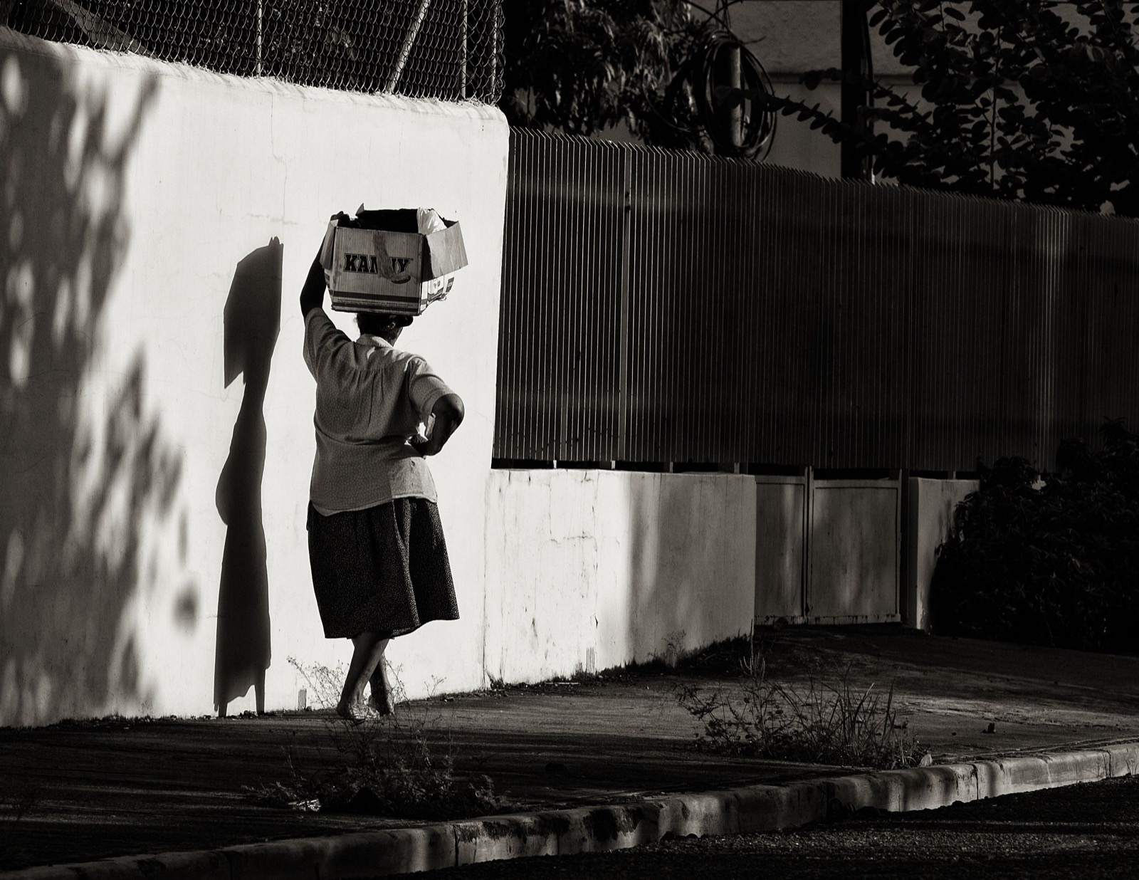 A man carries a box on his head along a sunlit wall, black and white